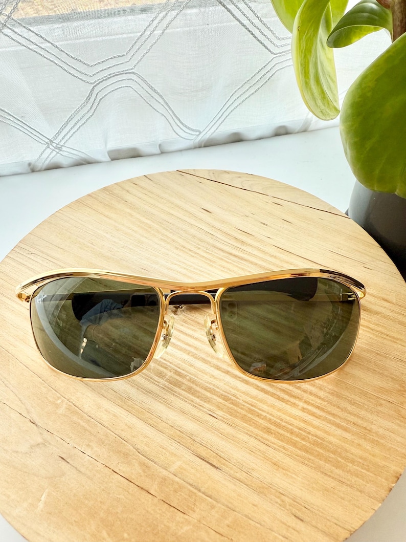 Peut inclure: Lunettes de soleil dor&eacute;es dot&eacute;es d'une barre frontale incurv&eacute;e et &eacute;l&eacute;gante ainsi que de verres rectangulaires teint&eacute;s en vert fonc&eacute;. La monture pr&eacute;sente une finition m&eacute;tallique polie, des plaquettes r&eacute;glables et des branches fines et lisses.