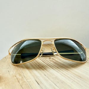 Peut inclure: Lunettes de soleil &agrave; monture en m&eacute;tal dor&eacute; avec un double pont et des verres teint&eacute;s vert fonc&eacute;. Le verre gauche affiche le logo Ray-Ban en blanc. Les fines branches dor&eacute;es sont termin&eacute;es par des embouts en plastique noir pour le confort.