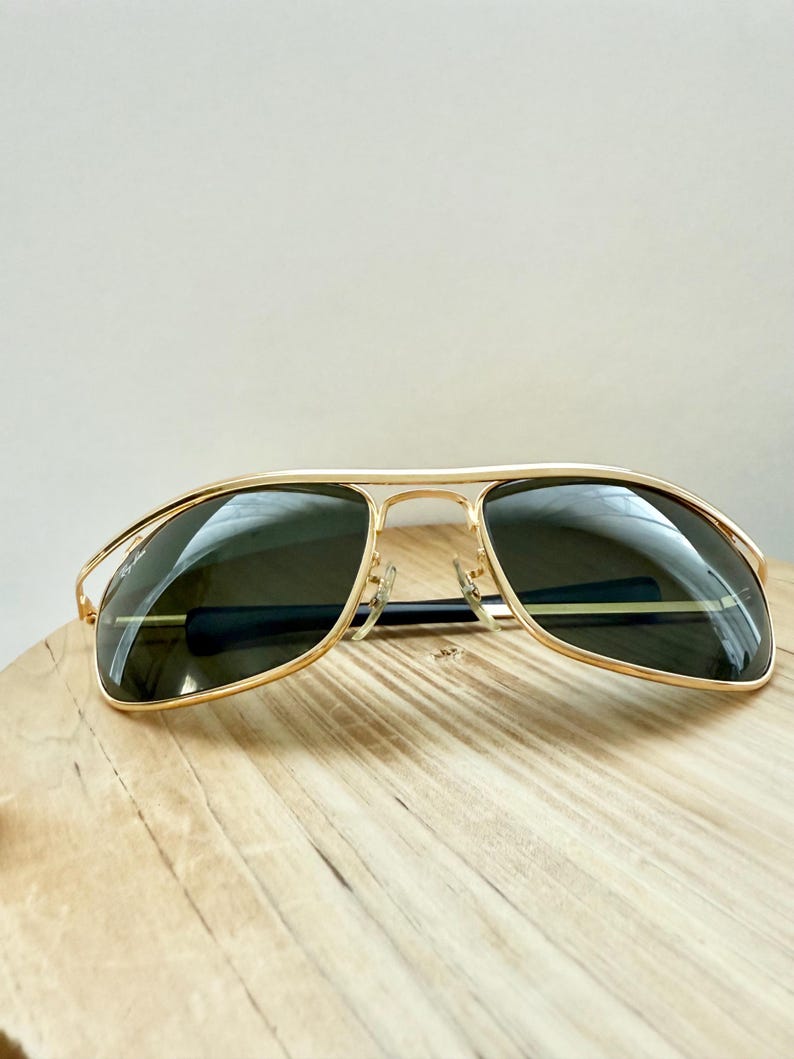 Peut inclure: Lunettes de soleil &agrave; monture en m&eacute;tal dor&eacute; avec un double pont et des verres teint&eacute;s vert fonc&eacute;. Le verre gauche affiche le logo Ray-Ban en blanc. Les fines branches dor&eacute;es sont termin&eacute;es par des embouts en plastique noir pour le confort.