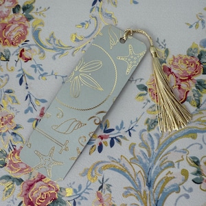 May include: A light blue bookmark with gold foil accents featuring a dragonfly, starfish, and seashell design. The bookmark has a gold tassel and is set against a floral patterned background.