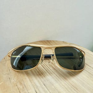 Peut inclure: Lunettes de soleil en m&eacute;tal dor&eacute; avec une barre frontale et des verres vert fonc&eacute;. Le verre gauche porte le logo Ray-Ban en blanc. La monture pr&eacute;sente un double pont avec des plaquettes de nez r&eacute;glables et des embouts de branches sombres.