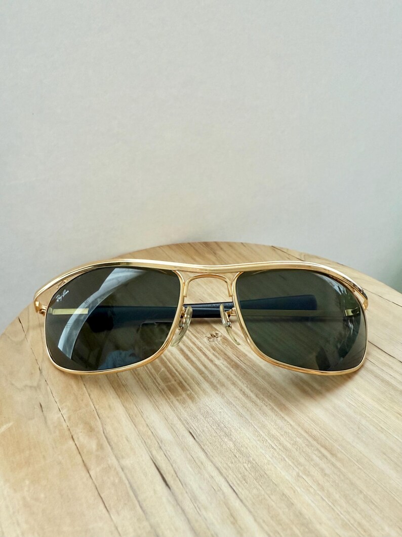 Peut inclure: Lunettes de soleil en m&eacute;tal dor&eacute; avec une barre frontale et des verres vert fonc&eacute;. Le verre gauche porte le logo Ray-Ban en blanc. La monture pr&eacute;sente un double pont avec des plaquettes de nez r&eacute;glables et des embouts de branches sombres.