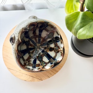 May include: A small, round, handmade ceramic bowl with an irregular, organic rim. The interior features a glossy glaze with a dark blue and brown grid pattern over a cream-colored base. The exterior is speckled with small dark dots on a light gray surface.