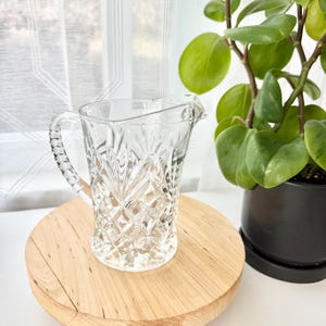 May include: A clear glass pitcher featuring a textured diamond and starburst pattern on the exterior body. The pitcher has a curved handle with a beaded design and a small pouring spout at the top rim, showcasing a classic pressed glass construction.
