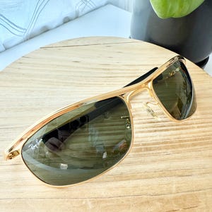 Peut inclure: Lunettes de soleil &agrave; monture en m&eacute;tal dor&eacute; avec un design &agrave; double pont et des verres teint&eacute;s vert fonc&eacute;. Le verre gauche affiche le logo blanc Ray-Ban dans le coin sup&eacute;rieur. Les lunettes incluent des plaquettes transparentes r&eacute;glables.