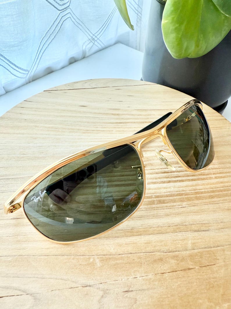 Peut inclure: Lunettes de soleil &agrave; monture en m&eacute;tal dor&eacute; avec un design &agrave; double pont et des verres teint&eacute;s vert fonc&eacute;. Le verre gauche affiche le logo blanc Ray-Ban dans le coin sup&eacute;rieur. Les lunettes incluent des plaquettes transparentes r&eacute;glables.