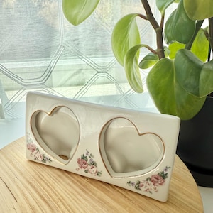 May include: A rectangular, off-white ceramic picture frame with two heart-shaped openings. The frame is decorated with gold trim and floral accents in pink, green, and white. The frame sits on a light wood surface.