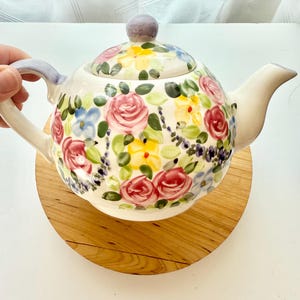 May include: A white ceramic teapot featuring a hand-painted floral pattern with pink roses, yellow blossoms, and blue accents. The teapot has a matching lid with a light purple knob, a curved handle with purple detailing, and a wide spout.