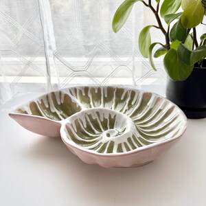 May include: A pink ceramic serving dish with a unique, nautilus-like design. The dish features three sections with a white and olive green pattern. The dish is on a white surface, with a plant in the background.