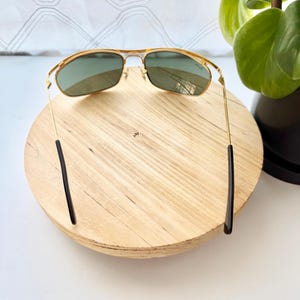 Peut inclure: Lunettes de soleil en m&eacute;tal dor&eacute; avec un design &agrave; double pont et des verres teint&eacute;s verts. Les fines montures m&eacute;talliques sont reli&eacute;es par une barre sup&eacute;rieure et les branches se terminent par des embouts en plastique noir. Logo Ray-Ban visible.