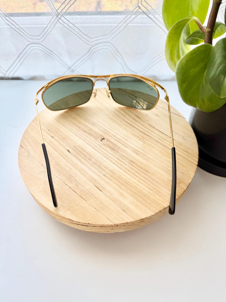 Peut inclure: Lunettes de soleil en m&eacute;tal dor&eacute; avec un design &agrave; double pont et des verres teint&eacute;s verts. Les fines montures m&eacute;talliques sont reli&eacute;es par une barre sup&eacute;rieure et les branches se terminent par des embouts en plastique noir. Logo Ray-Ban visible.