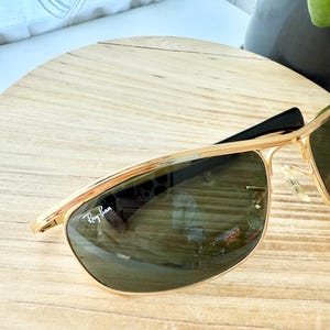 Peut inclure: Lunettes de soleil &agrave; monture en m&eacute;tal dor&eacute; avec un double pont et des verres vert fonc&eacute;. Le verre gauche pr&eacute;sente le logo Ray-Ban blanc imprim&eacute; dans le coin sup&eacute;rieur. Les branches sont noires et les plaquettes sont en plastique transparent.