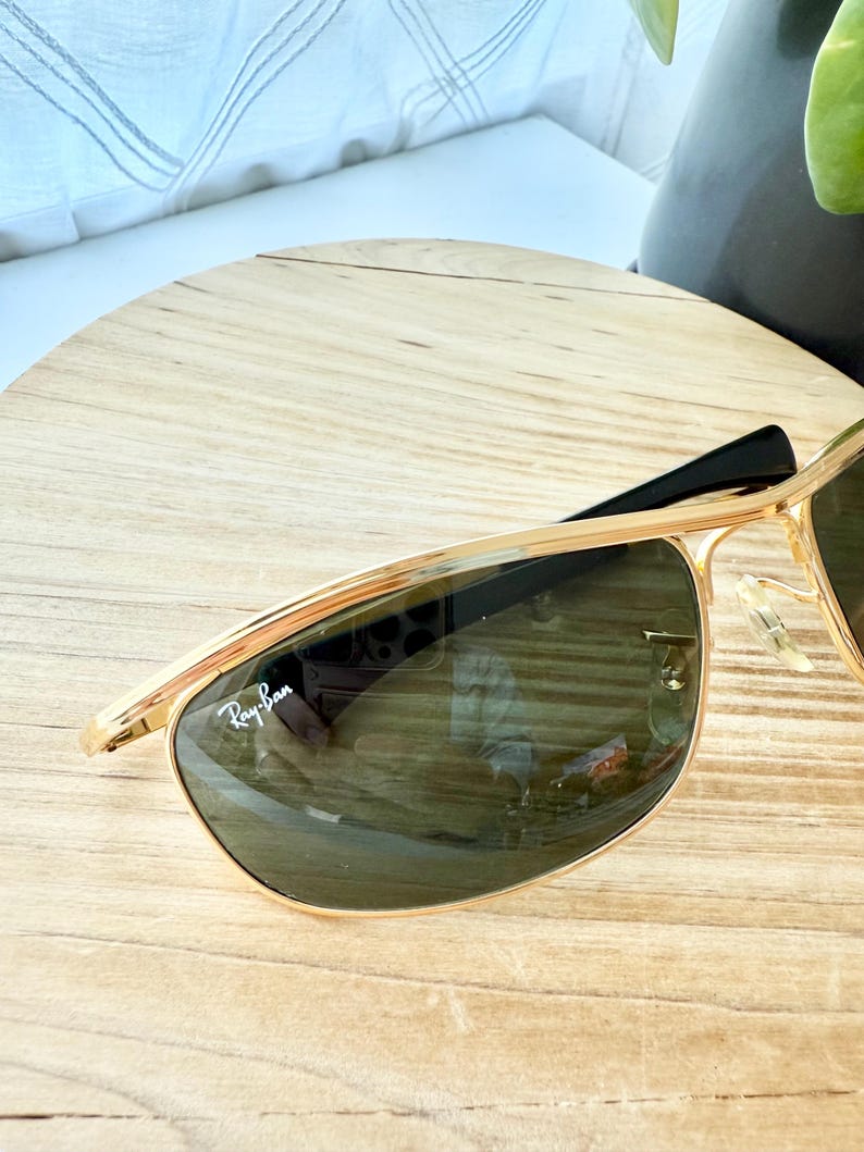 Peut inclure: Lunettes de soleil &agrave; monture en m&eacute;tal dor&eacute; avec un double pont et des verres vert fonc&eacute;. Le verre gauche pr&eacute;sente le logo Ray-Ban blanc imprim&eacute; dans le coin sup&eacute;rieur. Les branches sont noires et les plaquettes sont en plastique transparent.