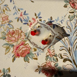May include: A pair of red teardrop earrings with gold-colored accents, displayed on a decorative clear holder. The earrings are set against a floral patterned background with pink and blue roses.