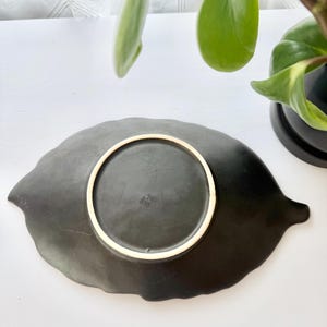 May include: A matte black ceramic dish shaped like a leaf, viewed from the underside. The base features a circular foot ring with a light-coloured rim and faint, embossed markings centred within the circle, showing a textured, dark surface.