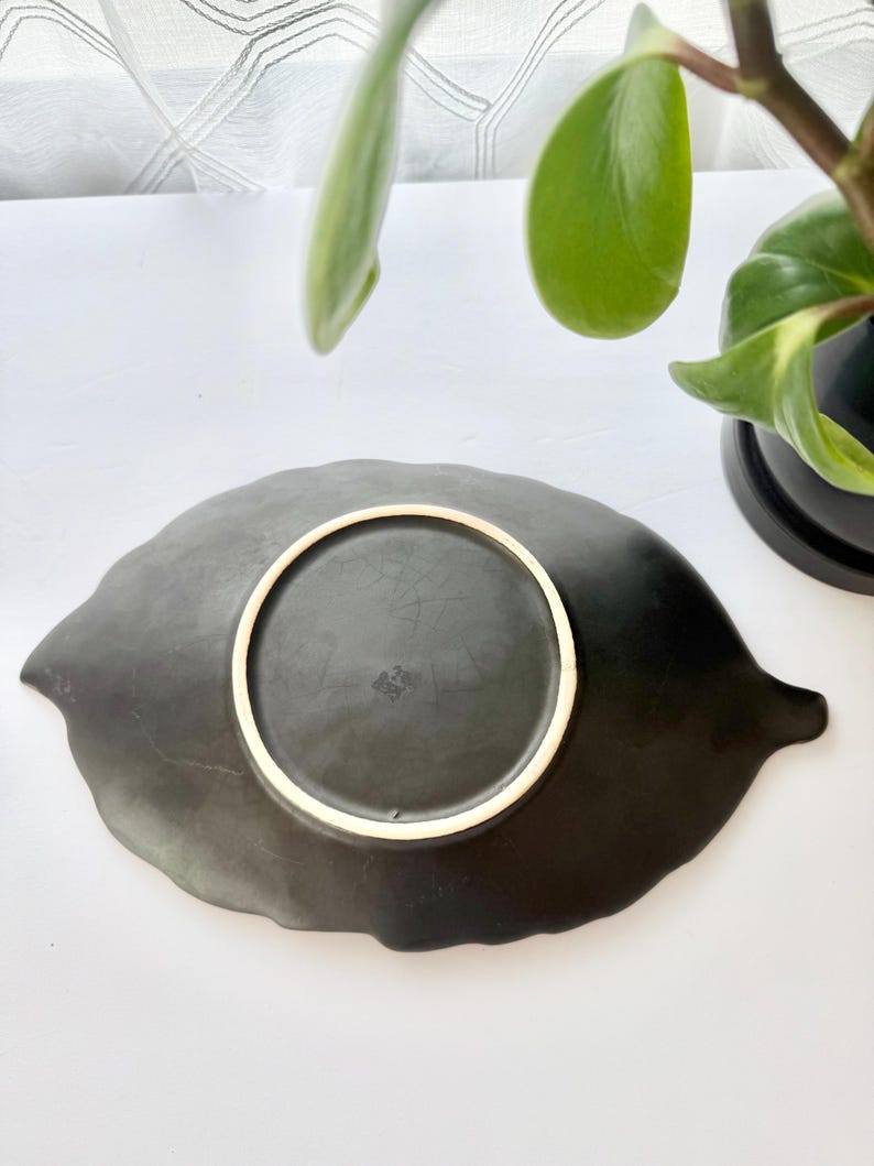 May include: A matte black ceramic dish shaped like a leaf, viewed from the underside. The base features a circular foot ring with a light-coloured rim and faint, embossed markings centred within the circle, showing a textured, dark surface.