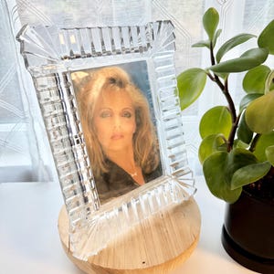May include: A clear, rectangular crystal picture frame with a portrait inside, resting on a round wooden base. The frame has a detailed, textured design. A potted plant is visible in the background.