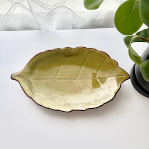 May include: A ceramic dish shaped like a leaf with a glossy finish. The dish features a gradient of light green to olive green colors with a dark brown rim. The surface has embossed vein details that mimic the natural structure of a leaf.