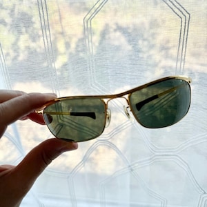 Peut inclure: Lunettes de soleil &agrave; monture en m&eacute;tal dor&eacute; avec des verres rectangulaires et un double pont. Les verres sont teint&eacute;s d'une nuance vert fonc&eacute;, et les branches fines en m&eacute;tal sont dot&eacute;es d'embouts en plastique noir pour plus de confort.