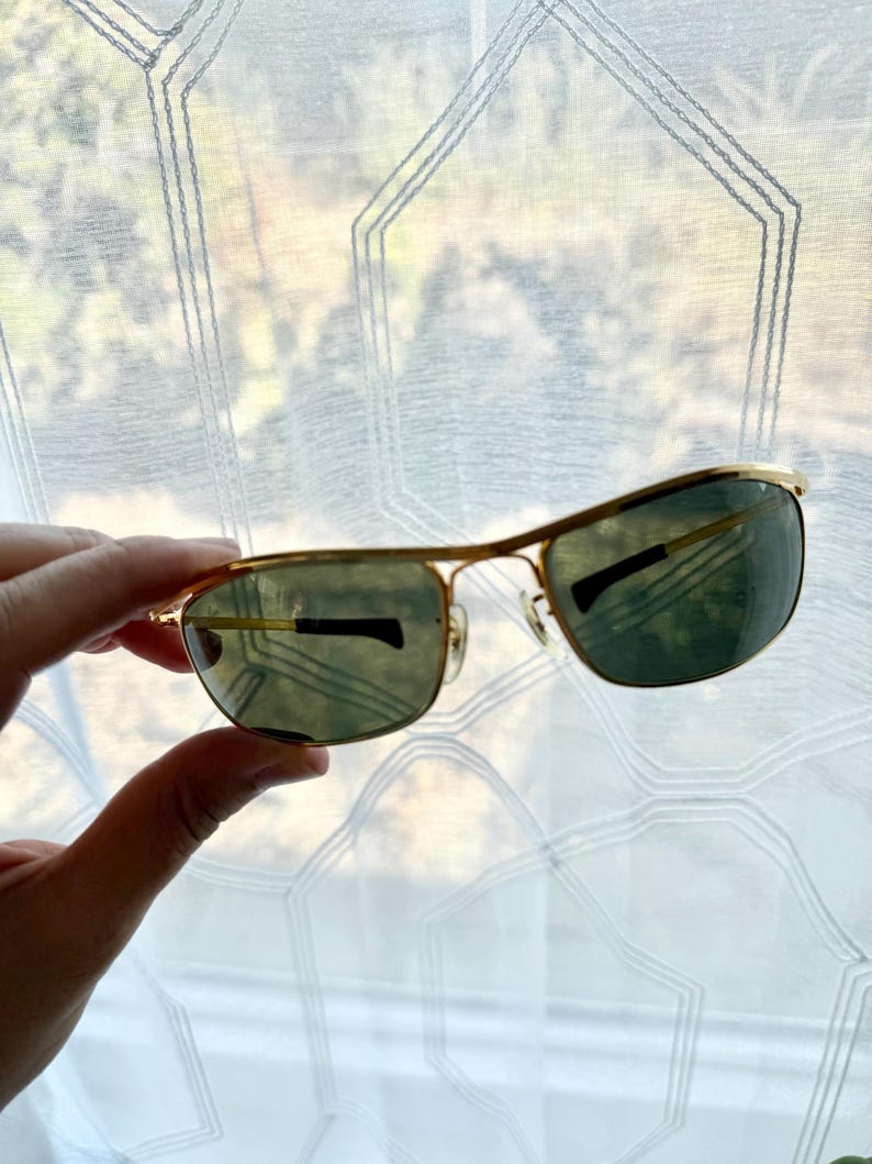 Peut inclure: Lunettes de soleil &agrave; monture en m&eacute;tal dor&eacute; avec des verres rectangulaires et un double pont. Les verres sont teint&eacute;s d'une nuance vert fonc&eacute;, et les branches fines en m&eacute;tal sont dot&eacute;es d'embouts en plastique noir pour plus de confort.
