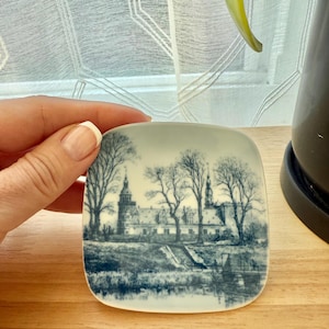 May include: Square ceramic trinket dish with a blue and white illustration of a castle and trees. The dish has a slightly curved edge and a glossy finish. The artwork depicts a landscape scene.