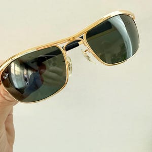 Peut inclure: Lunettes de soleil en m&eacute;tal dor&eacute; avec un design &agrave; double pont et des verres vert fonc&eacute;. Le verre gauche affiche le logo Ray-Ban en &eacute;criture blanche. La monture est dot&eacute;e de plaquettes de nez r&eacute;glables et d'embouts de branches noirs.