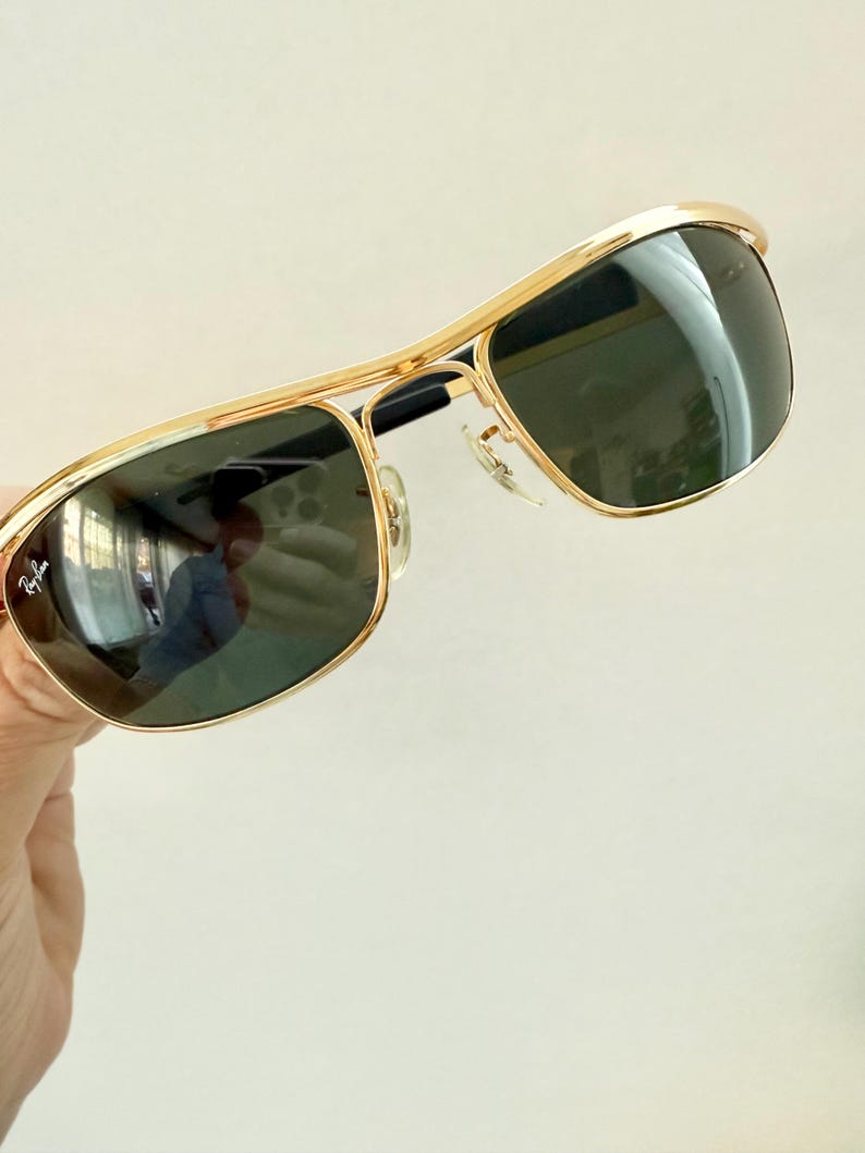 Peut inclure: Lunettes de soleil en m&eacute;tal dor&eacute; avec un design &agrave; double pont et des verres vert fonc&eacute;. Le verre gauche affiche le logo Ray-Ban en &eacute;criture blanche. La monture est dot&eacute;e de plaquettes de nez r&eacute;glables et d'embouts de branches noirs.