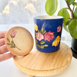 May include: A cylindrical blue ceramic container featuring hand-painted pink and yellow roses with green leaves. A matching circular wooden lid is held to the side, decorated with a single painted pink rosebud and green stem on a light wood grain.