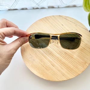 Peut inclure: Lunettes de soleil &agrave; monture en m&eacute;tal dor&eacute; avec des verres vert fonc&eacute;. Le verre gauche porte le logo Ray-Ban en blanc. Les lunettes pr&eacute;sentent un double pont, des branches fines en m&eacute;tal dor&eacute; et des embouts noirs. Les plaquettes sont transparentes.