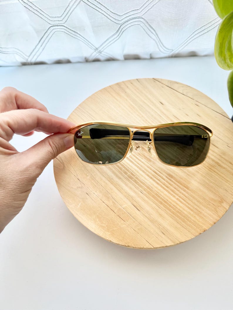 Peut inclure: Lunettes de soleil &agrave; monture en m&eacute;tal dor&eacute; avec des verres vert fonc&eacute;. Le verre gauche porte le logo Ray-Ban en blanc. Les lunettes pr&eacute;sentent un double pont, des branches fines en m&eacute;tal dor&eacute; et des embouts noirs. Les plaquettes sont transparentes.