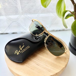 Peut inclure: Lunettes de soleil &agrave; monture en m&eacute;tal dor&eacute; avec verres vert fonc&eacute; pos&eacute;es sur un &eacute;tui noir en similicuir. L'&eacute;tui porte l'inscription dor&eacute;e Ray-Ban SUNGLASSES by BAUSCH & LOMB. Les lunettes pr&eacute;sentent un design &agrave; double pont.