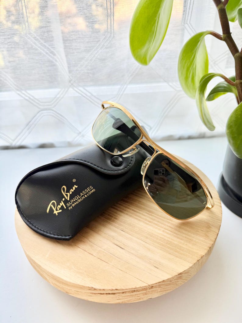 Peut inclure: Lunettes de soleil &agrave; monture en m&eacute;tal dor&eacute; avec verres vert fonc&eacute; pos&eacute;es sur un &eacute;tui noir en similicuir. L'&eacute;tui porte l'inscription dor&eacute;e Ray-Ban SUNGLASSES by BAUSCH & LOMB. Les lunettes pr&eacute;sentent un design &agrave; double pont.