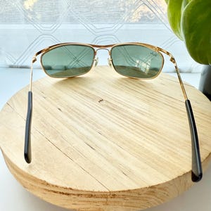 Peut inclure: Lunettes de soleil en m&eacute;tal dor&eacute; avec des verres rectangulaires teint&eacute;s en vert et des embouts de branches noirs. Le logo Ray-Ban est visible dans le coin sup&eacute;rieur du verre droit. La monture pr&eacute;sente un double pont et des plaquettes.
