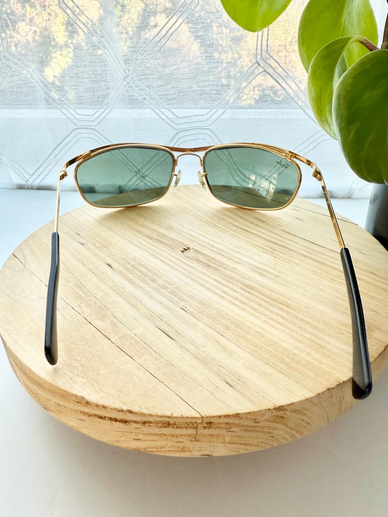 Peut inclure: Lunettes de soleil en m&eacute;tal dor&eacute; avec des verres rectangulaires teint&eacute;s en vert et des embouts de branches noirs. Le logo Ray-Ban est visible dans le coin sup&eacute;rieur du verre droit. La monture pr&eacute;sente un double pont et des plaquettes.