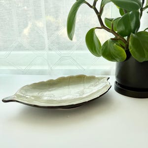 May include: A ceramic dish shaped like a leaf with a glossy, light green glaze and a dark brown rim. The surface features embossed vein details that mimic the natural structure of a leaf, creating a textured appearance across the shallow bowl.