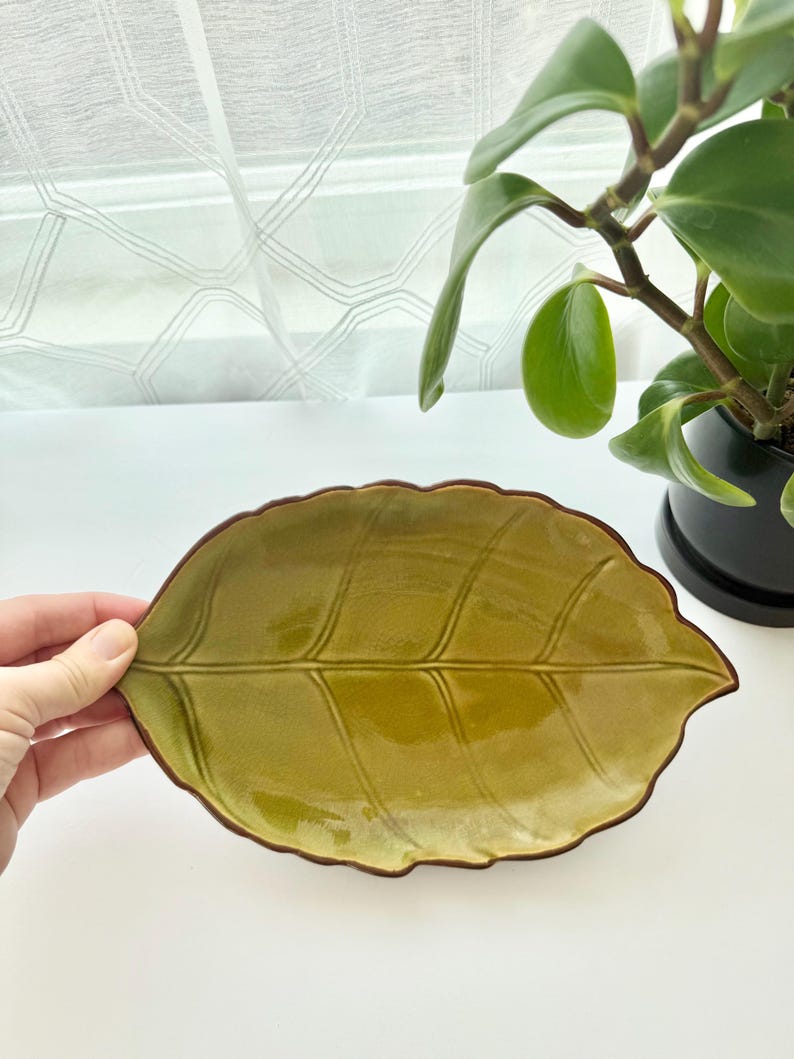 May include: An oval-shaped ceramic dish designed to resemble a leaf, featuring a glossy olive green glaze with a dark brown rim. The surface of the dish is textured with embossed veins that mimic the natural structure of a leaf, creating a detailed botanical appearance.