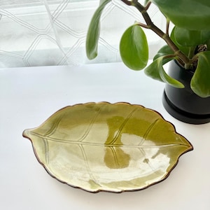 May include: A ceramic dish shaped like a leaf with a glossy, light green glaze and a dark brown rim. The surface features embossed vein details that mimic the natural structure of a leaf, creating a textured appearance on the shallow, oval-shaped tray.