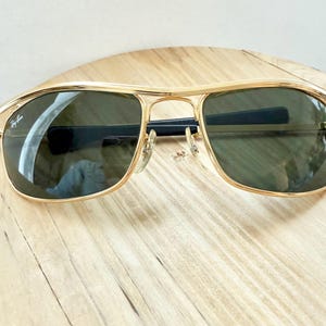 Peut inclure: Lunettes de soleil &agrave; monture en m&eacute;tal dor&eacute; avec un design &agrave; double pont et des verres teint&eacute;s vert fonc&eacute;. Le verre gauche pr&eacute;sente le logo Ray-Ban en &eacute;criture blanche. Les branches sont noires et les plaquettes sont transparentes.