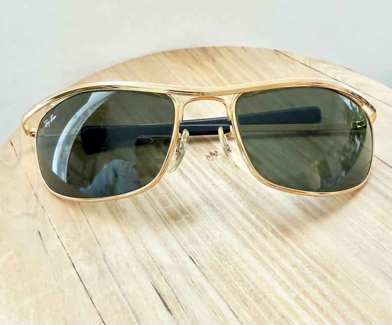 Peut inclure: Lunettes de soleil &agrave; monture en m&eacute;tal dor&eacute; avec un design &agrave; double pont et des verres teint&eacute;s vert fonc&eacute;. Le verre gauche pr&eacute;sente le logo Ray-Ban en &eacute;criture blanche. Les branches sont noires et les plaquettes sont transparentes.