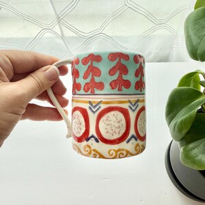 May include: A ceramic mug featuring a light blue upper band with red leaf-like patterns, followed by a middle section with red circles containing speckled centers, and a lower band with yellow scrollwork designs on a white background.