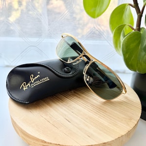 Peut inclure: Lunettes de soleil &agrave; monture en m&eacute;tal dor&eacute; avec verres teint&eacute;s verts et un double pont. Les lunettes sont plac&eacute;es &agrave; c&ocirc;t&eacute; d'un &eacute;tui de protection en similicuir noir portant le texte imprim&eacute; en dor&eacute; Ray-Ban Sunglasses by Bausch & Lomb.