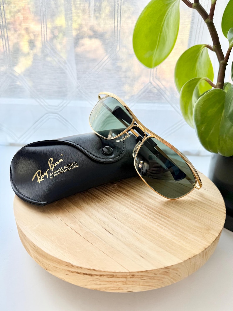 Peut inclure: Lunettes de soleil &agrave; monture en m&eacute;tal dor&eacute; avec verres teint&eacute;s verts et un double pont. Les lunettes sont plac&eacute;es &agrave; c&ocirc;t&eacute; d'un &eacute;tui de protection en similicuir noir portant le texte imprim&eacute; en dor&eacute; Ray-Ban Sunglasses by Bausch & Lomb.