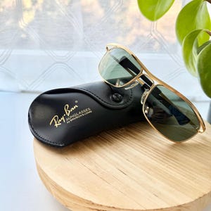 Peut inclure: Lunettes de soleil &agrave; monture en m&eacute;tal dor&eacute; avec des verres vert fonc&eacute; pos&eacute;es sur un &eacute;tui en similicuir noir. L'&eacute;tui porte le texte imprim&eacute; en dor&eacute; Ray-Ban SUNGLASSES by BAUSCH & LOMB. Les lunettes pr&eacute;sentent un double pont.
