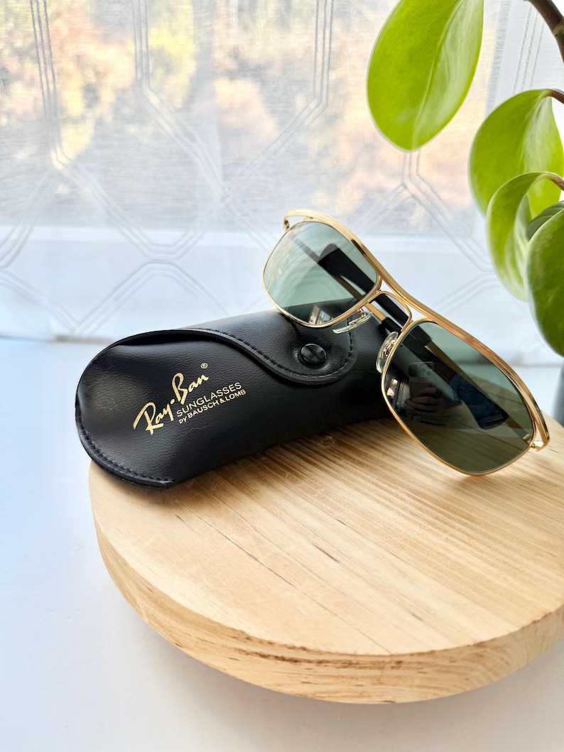 Peut inclure: Lunettes de soleil &agrave; monture en m&eacute;tal dor&eacute; avec des verres vert fonc&eacute; pos&eacute;es sur un &eacute;tui en similicuir noir. L'&eacute;tui porte le texte imprim&eacute; en dor&eacute; Ray-Ban SUNGLASSES by BAUSCH & LOMB. Les lunettes pr&eacute;sentent un double pont.