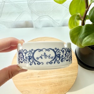 May include: A white ceramic ashtray featuring a blue decorative scrollwork pattern and the word "orbis" printed in a cursive script on the front. The circular ashtray has two notches on the rim for resting items and a smooth, glossy finish.
