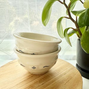 May include: Two off-white ceramic bowls stacked on top of each other. Each bowl has a slightly flared rim and is decorated with small blue floral and curved line designs. The bowls sit on a round, light-colored wooden surface.