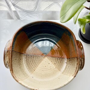 May include: A handmade ceramic bowl with a unique design. The bowl features a combination of colors, including brown, beige, blue, and orange. It has a round shape with two handles on the sides. The interior has a geometric pattern.