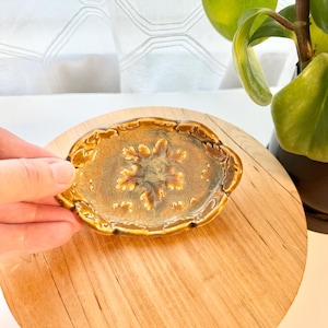 May include: An oval ceramic dish featuring a glossy brown and tan glaze with a raised, textured oak leaf motif in the center. The edges of the dish are scalloped and finished with a darker brown trim, creating a decorative border around the design.