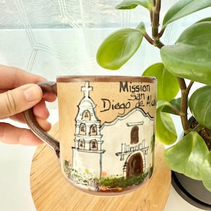 May include: A tan ceramic mug featuring a hand-painted illustration of Mission San Diego de Alcala with a bell tower and arched doorway. The text Mission San Diego de Alcala is written in black ink on the upper right side of the mug surface.