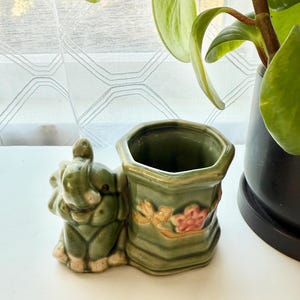 May include: A ceramic planter featuring a sculpted green elephant standing beside a hexagonal vessel. The vessel is decorated with a raised, painted branch with a pink flower. The entire piece has a glossy, sage green glaze finish.